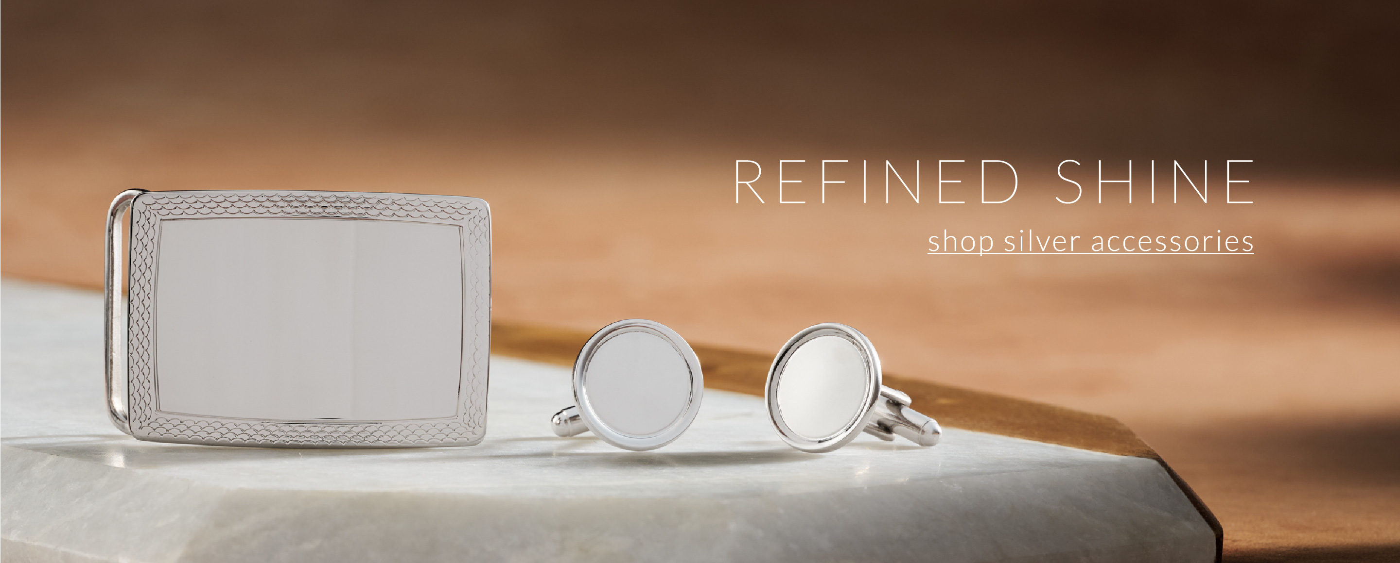 Shop Silver Accessories. A silver belt buckle and silver cufflinks on a marble slab.
