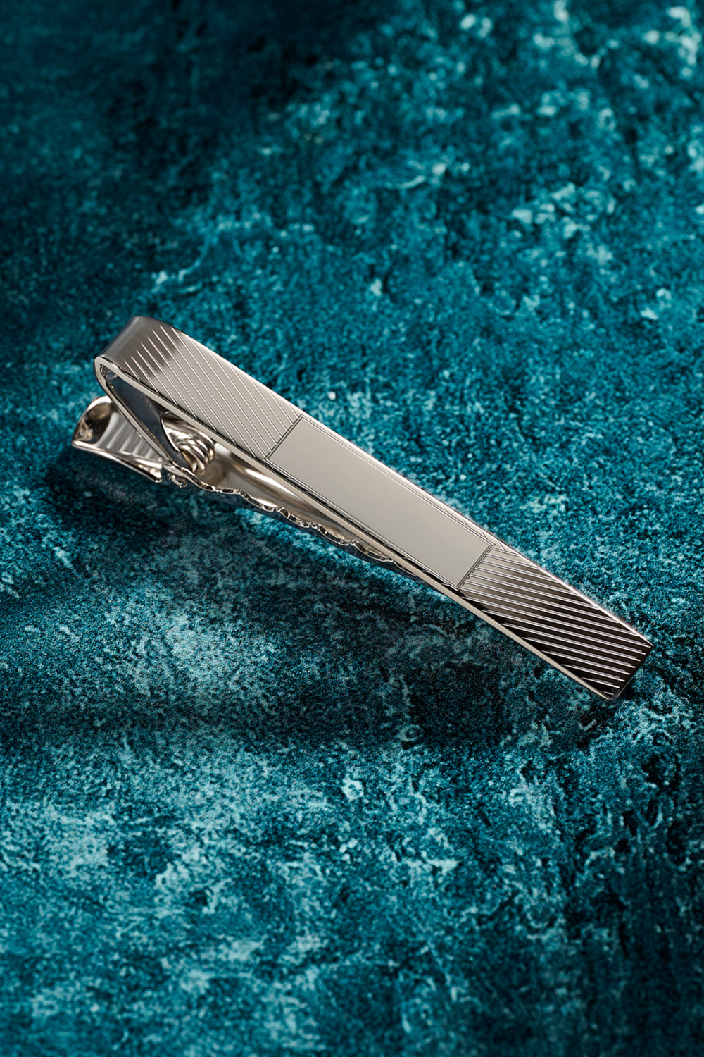 Silver engine turned tie bar on a textured blue background.