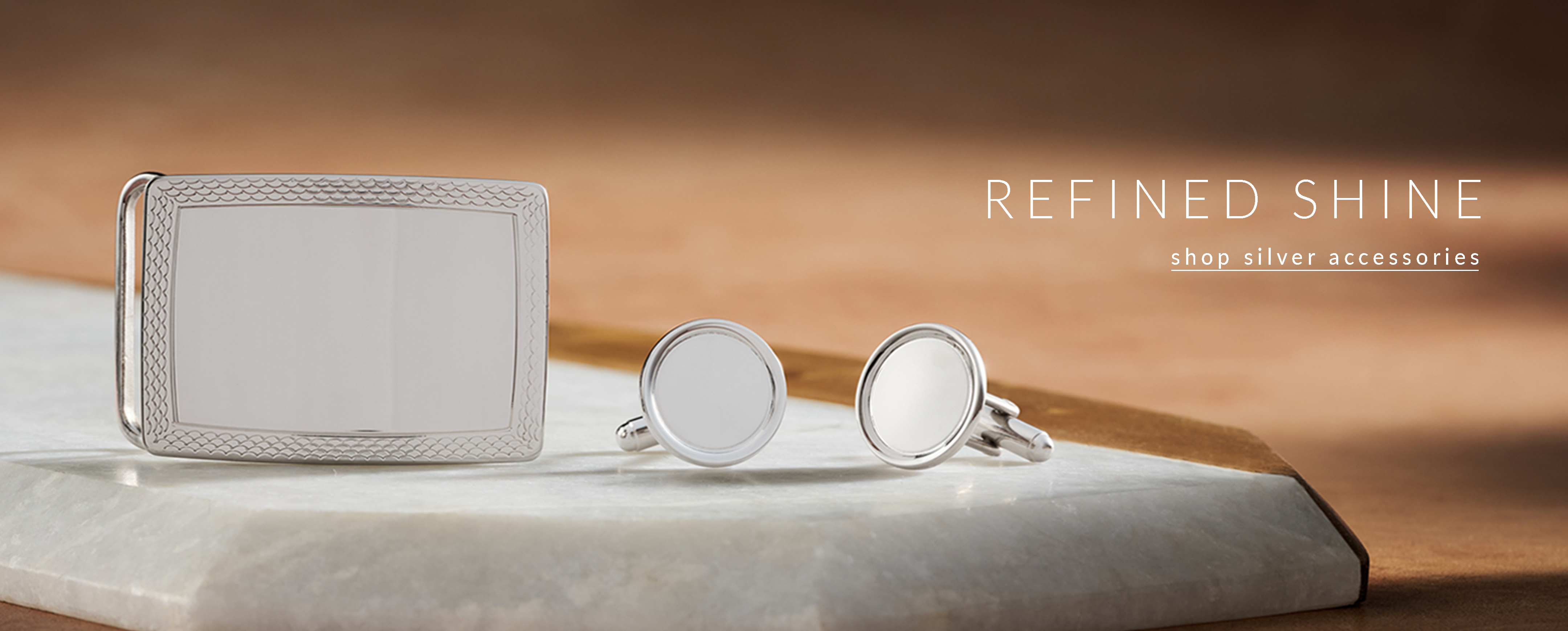 Shop Silver Accessories. A silver belt buckle and silver cufflinks on a marble slab.
